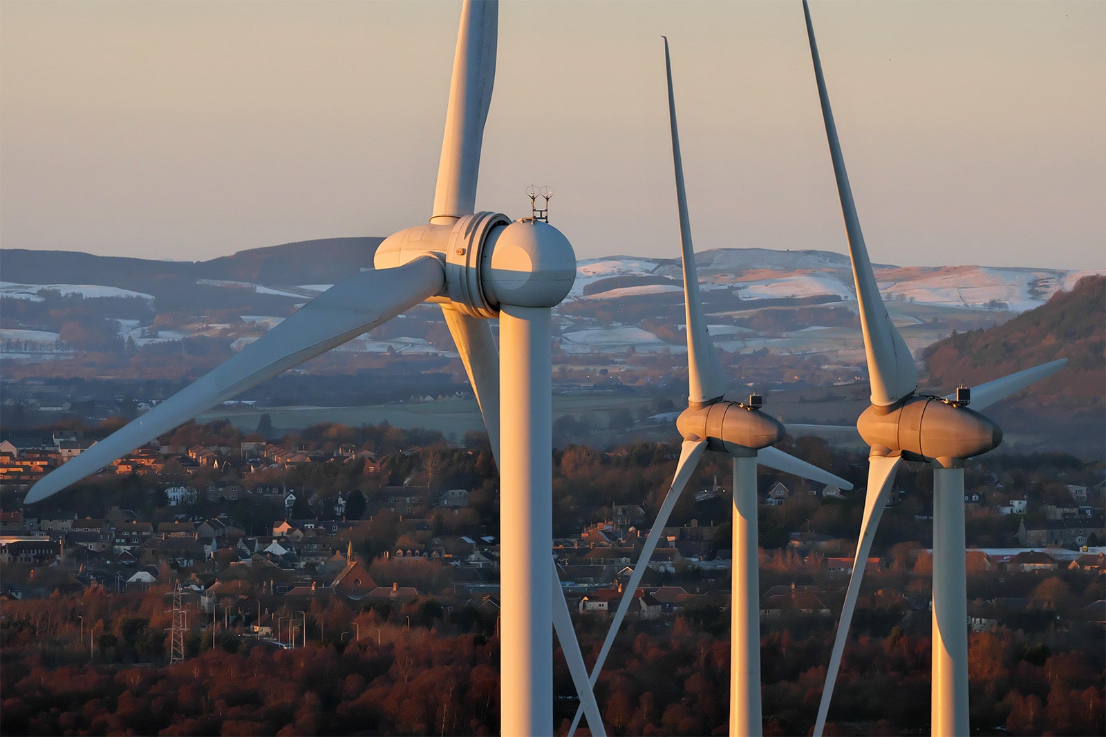 Three wind turbines rise above a Scottish town, set against a picturesque landscape of hills.