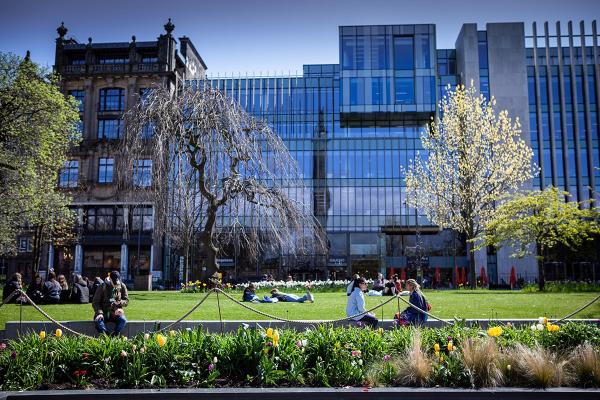 A park in a city on a spring day, with flowers in the foreground, people relaxing on a lawn and modern office buildings behind