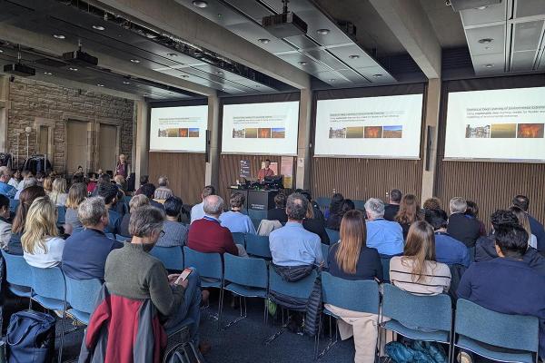 View from the back of a large meeting room with rows of people watching a presentation