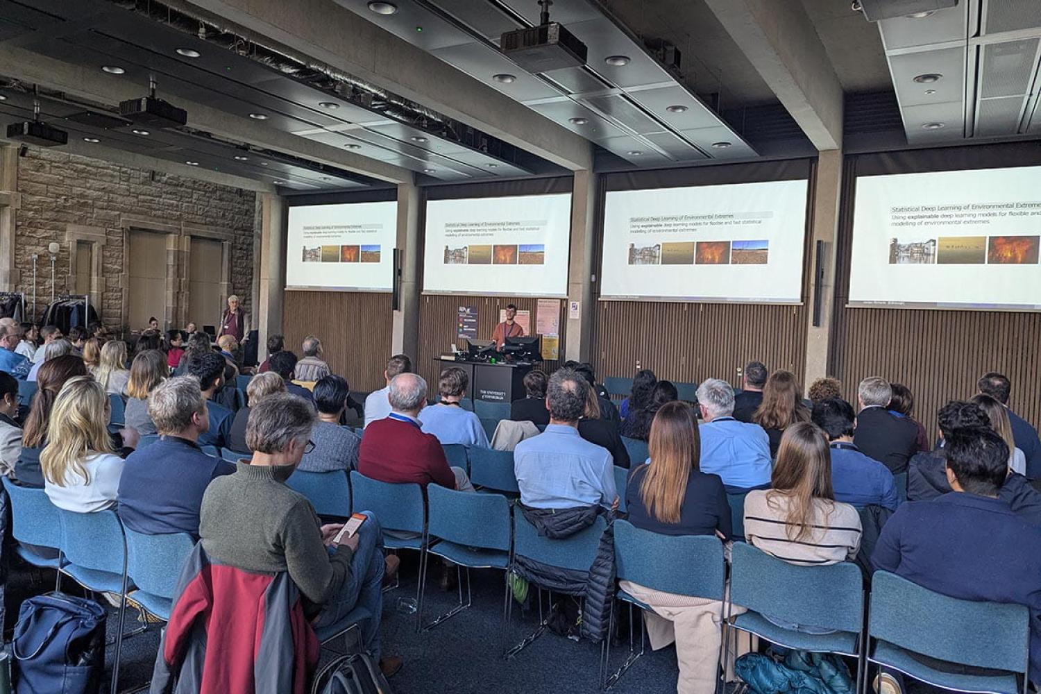 View from the back of a large meeting room with rows of people watching a presentation